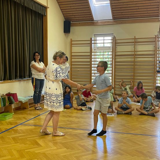 Eine Frau und ein Junge schütteln sich die Hände in einer Turnhalle, während Kinder auf dem Boden sitzen.