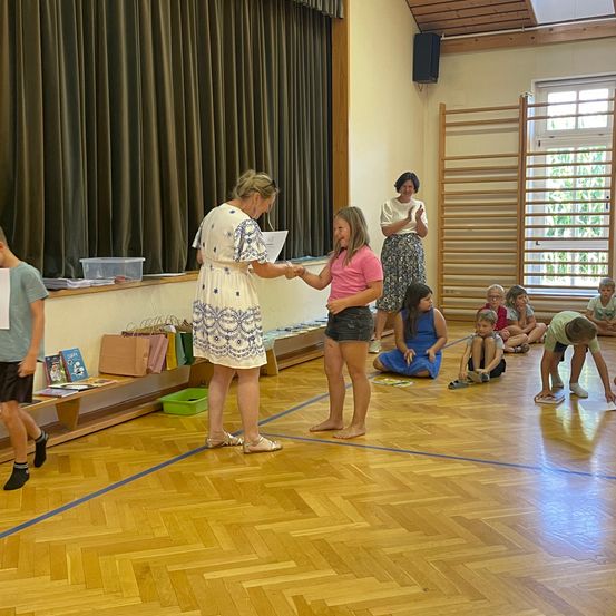 Eine Frau überreicht einer jungen Mädchen in einem rosa T-Shirt ein Zertifikat in einer Klassenzimmerumgebung. Andere Kinder sitzen und stehen herum, und eine Frau klatscht im Hintergrund.