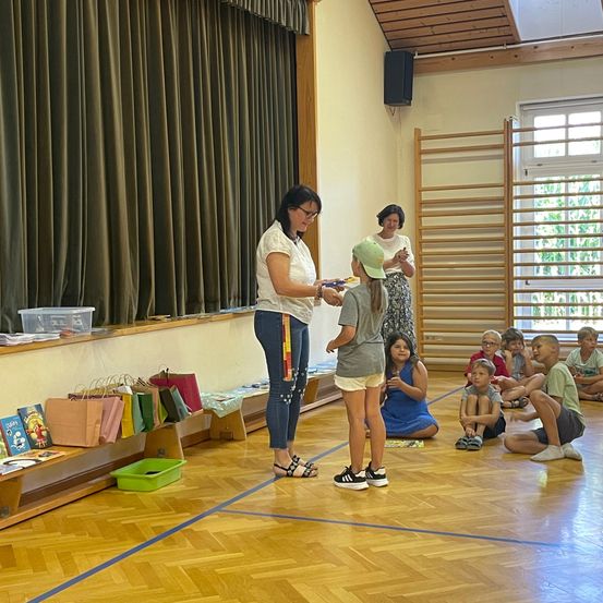 Eine Frau überreicht einem Mädchen ein Buch in einem Klassenzimmer, in dem andere Kinder auf dem Boden sitzen.