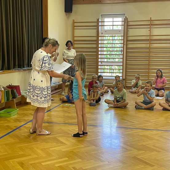 Eine Frau überreicht einer jungen Mädchen in einem blauen Kleid ein Zertifikat, während Kinder im Kreis auf dem Boden sitzen und zusehen.