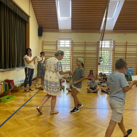Eine Gruppe von Kindern und Erwachsenen versammelt sich in einem Raum, einige sitzen auf dem Boden, andere stehen. Eine Frau in einem weißen Kleid klatscht.