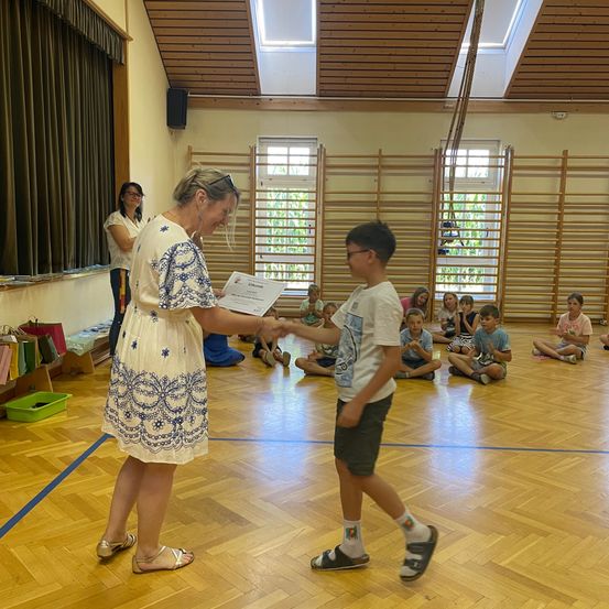 Eine Frau in einem Blumenkleid überreicht einem jungen Jungen ein Zertifikat in einer Klassenzimmerumgebung, mit Kindern, die im Hintergrund auf dem Boden sitzen.