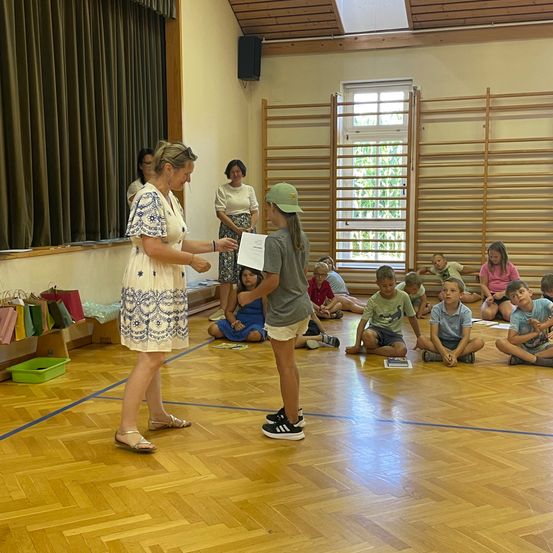 Eine Frau in einem weißen Kleid überreicht einem jungen Mädchen in einer Turnhalle ein Zertifikat, während Kinder auf dem Boden sitzen.