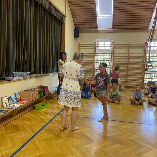 Eine Frau überreicht einem Mädchen eine Urkunde in einem Raum, in dem Kinder auf dem Boden sitzen. Eine Frau steht hinter ihnen, und ein Regal mit Büchern und Taschen befindet sich in der Nähe.