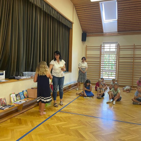 Zwei Frauen und mehrere Kinder befinden sich in einem Raum mit Holzböden. Eine Frau hält ein Buch und spricht mit einem jungen Mädchen. Andere Kinder sitzen auf dem Boden, einige lesen Bücher.
