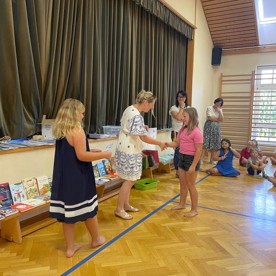Zwei Mädchen erhalten ein Buch von einer Frau in einer Klassenzimmerumgebung, in der andere Kinder auf dem Boden sitzen.