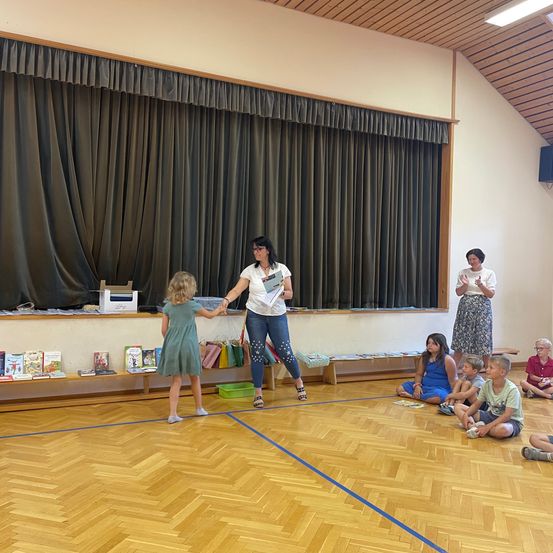 Eine Frau, die ein Buch in der Hand hält, steht vor einer Gruppe von Kindern, die auf dem Boden in einem Raum mit Holzböden und einer Bühne mit Vorhängen sitzen.