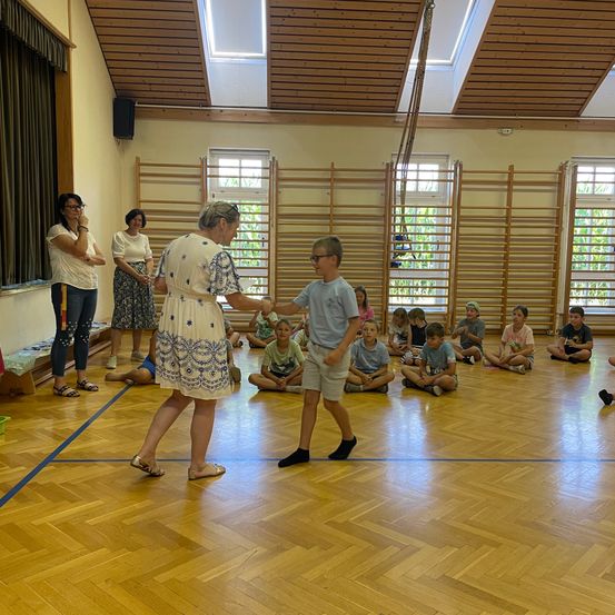 Eine Frau und ein Junge schütteln sich die Hände in einer Turnhalle, während mehrere Kinder auf dem Boden sitzen und zusehen.