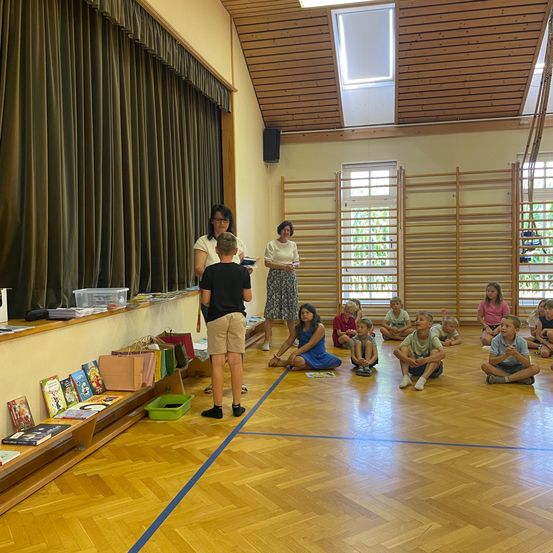Eine Gruppe von Kindern und zwei Erwachsenen befinden sich in einem Klassenzimmer. Die Kinder sitzen auf dem Boden, während die Erwachsenen in der Nähe eines Bücherregals stehen.