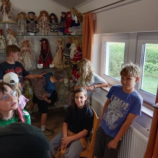 Eine Gruppe von Kindern versammelt sich um eine Puppenausstellung in einem Raum mit einem Fenster, das ein Grasfeld draußen zeigt.