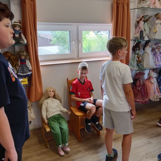 Drei Jungen stehen in einem Raum mit einer Puppe auf einem Stuhl. Der Junge im roten Trikot sitzt mit verschränkten Armen auf einem Stuhl.