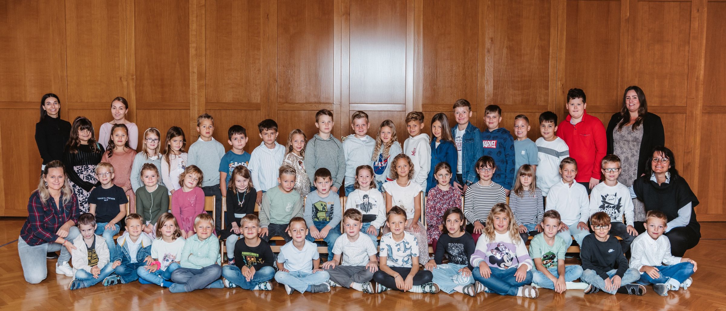 Eine Gruppe von Kindern und Jugendlichen, einige stehend und einige sitzend, posiert für ein Foto in einem Raum mit Holzwänden und -böden.