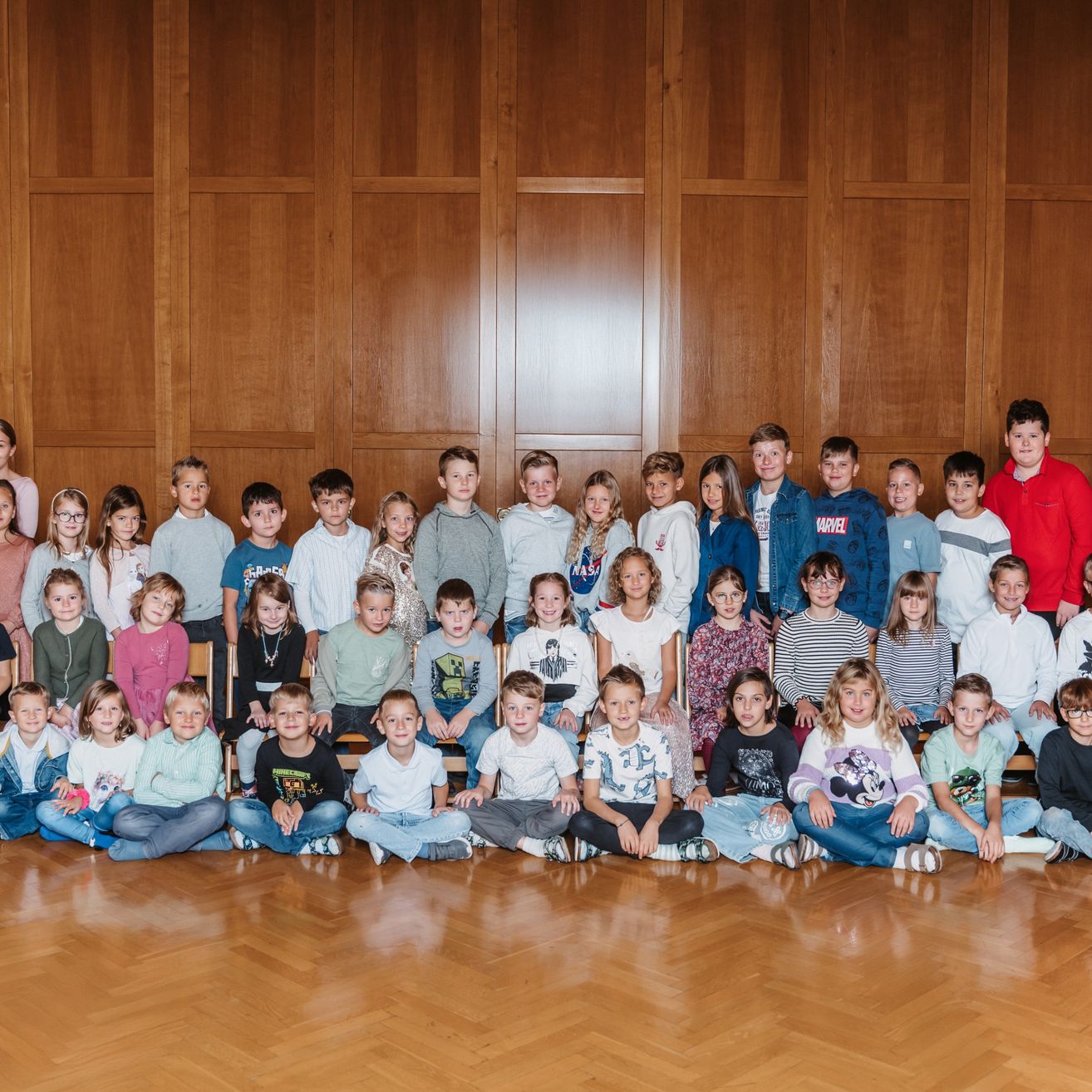 Eine Gruppe von Kindern, sowohl Jungen als auch Mädchen, sitzen und stehen in einer Reihe, wahrscheinlich in einem Klassenzimmer. Sie tragen alle verschiedene Kleidungsstücke und einige von ihnen tragen Brillen. Der Raum hat Holzwände und einen Holzboden.