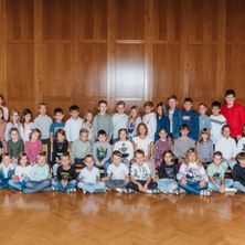 Eine Gruppe von Kindern, sowohl Jungen als auch Mädchen, sitzen und stehen in einer Reihe, wahrscheinlich in einem Klassenzimmer. Sie tragen alle verschiedene Kleidungsstücke und einige von ihnen tragen Brillen. Der Raum hat Holzwände und einen Holzboden.