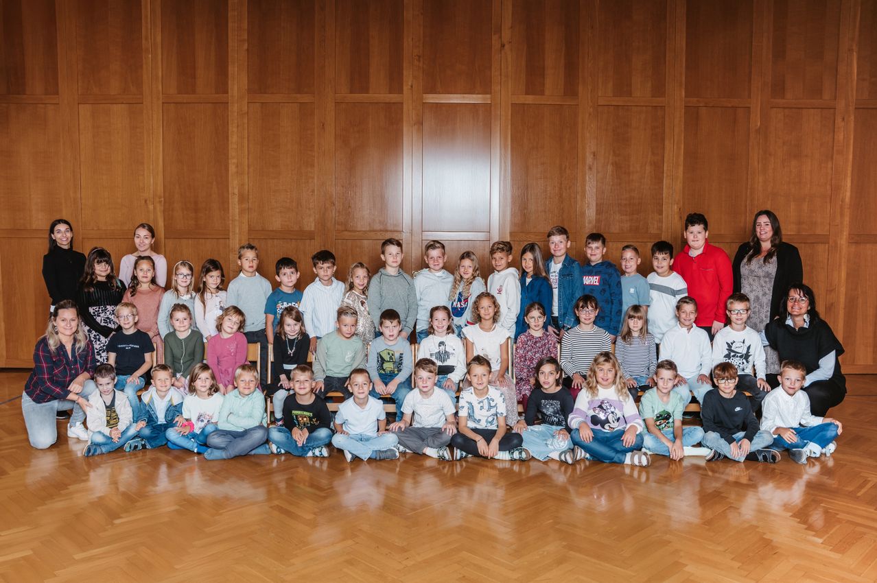 Eine Gruppe von Kindern, sowohl Jungen als auch Mädchen, sitzen und stehen in einer Reihe, wahrscheinlich in einem Klassenzimmer. Sie tragen alle verschiedene Kleidungsstücke und einige von ihnen tragen Brillen. Der Raum hat Holzwände und einen Holzboden.
