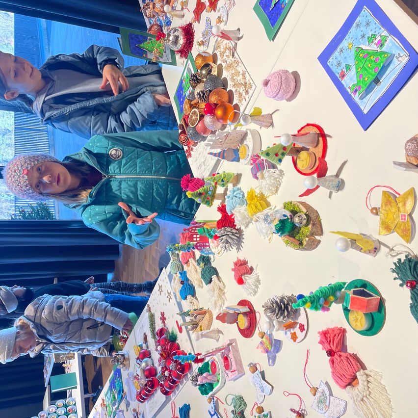 Drei Kinder stehen vor einem Tisch, der mit verschiedenen handgefertigten Weihnachtsdekorationen bedeckt ist. Der Tisch hat eine weiße Oberfläche mit bunten Ornamenten und Kunstwerken, einschließlich Ornamente, einem kleinen Weihnachtsbaum und einem kleinen Haus.