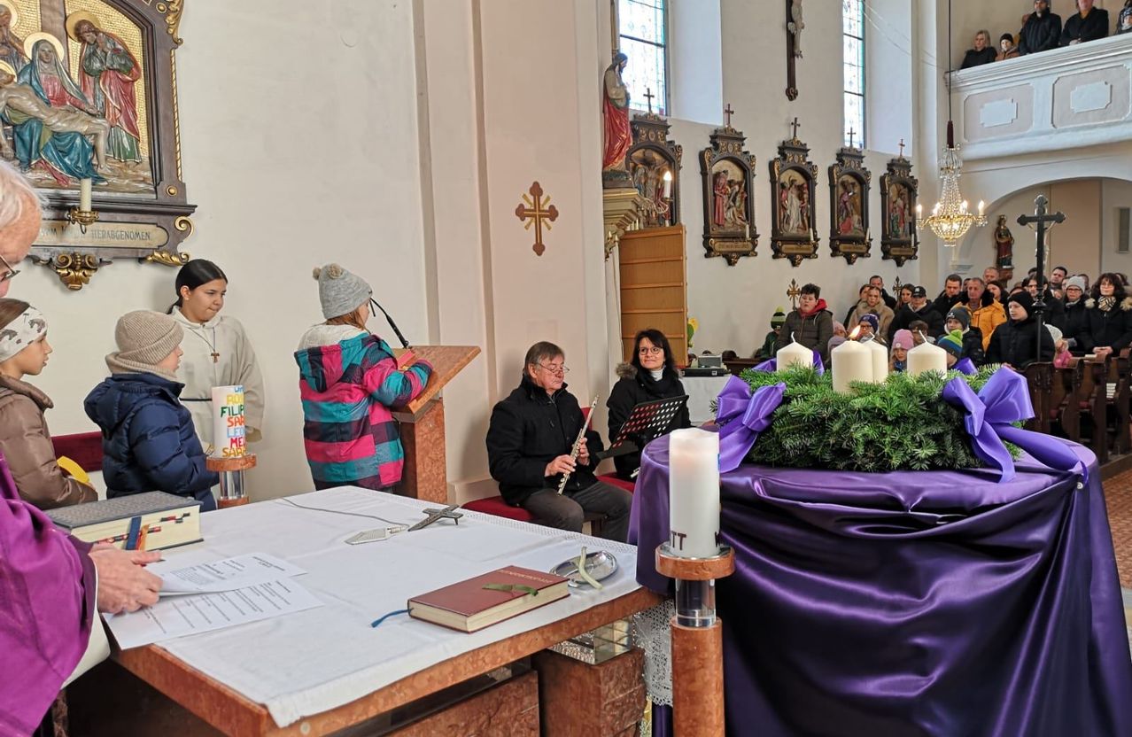 Eine Gruppe von Menschen in einer Kirche, wobei eine Person eine Flöte spielt. Es gibt Kerzen, ein Buch und ein Kreuz auf einem Tisch.