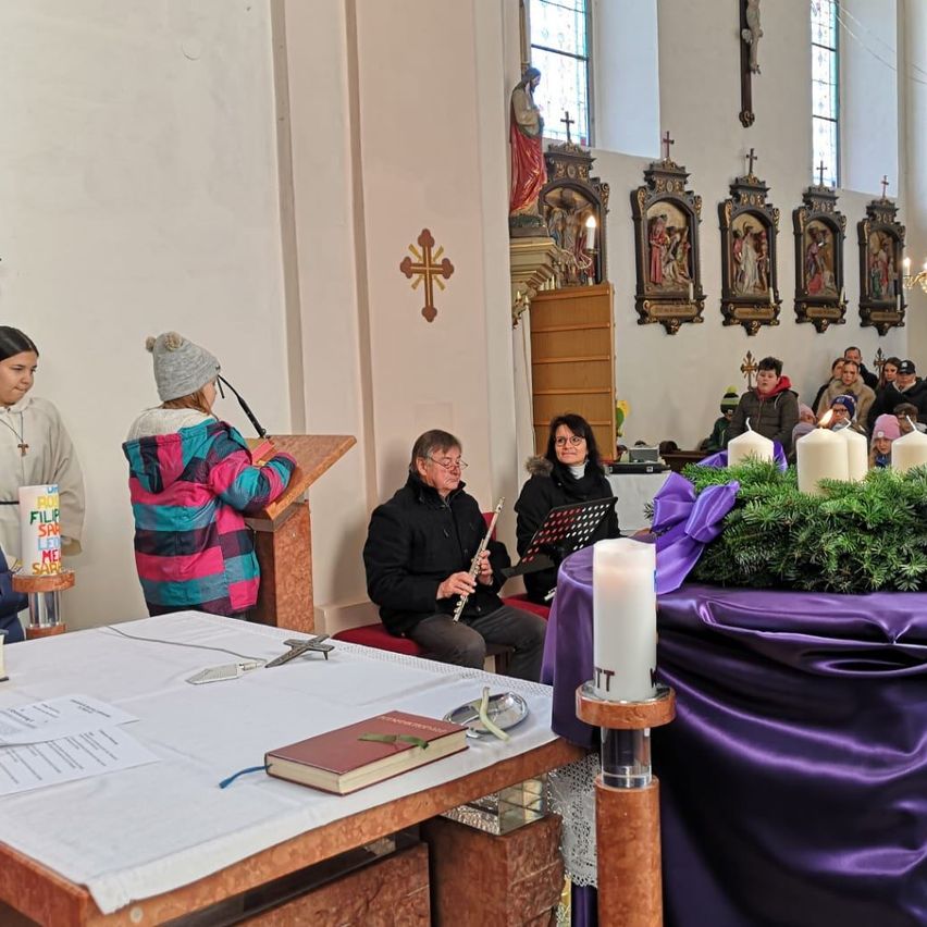 Eine Gruppe von Menschen in einer Kirche, wobei eine Person eine Flöte spielt. Es gibt Kerzen, ein Buch und ein Kreuz auf einem Tisch.