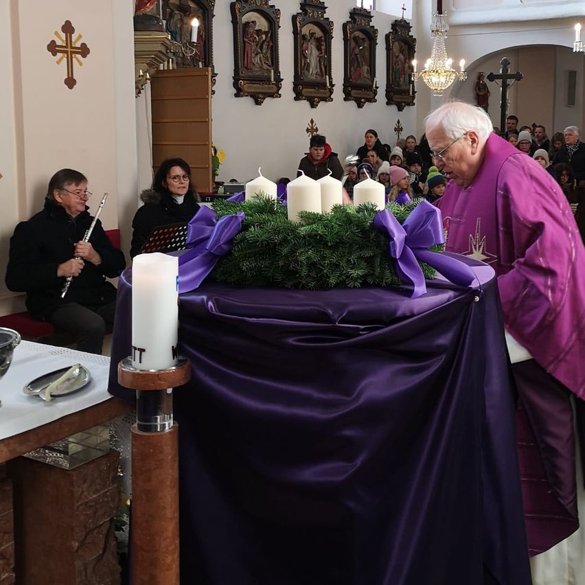 Ein Priester zündet Kerzen auf einem Tisch an, der mit einem Kranz geschmückt ist, während ein Mann in einer Kirche Flöte spielt.