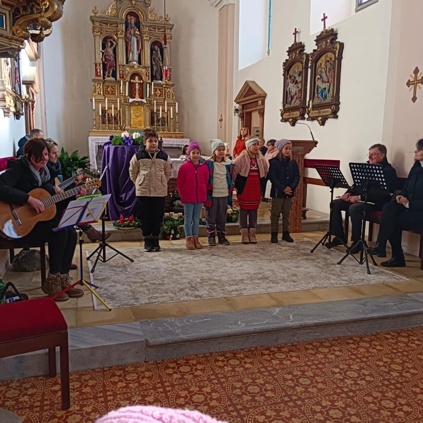 Eine Gruppe von Kindern steht vor einer Bühne in einer Kirche, begleitet von zwei Musikern, die Gitarre spielen und Noten lesen.