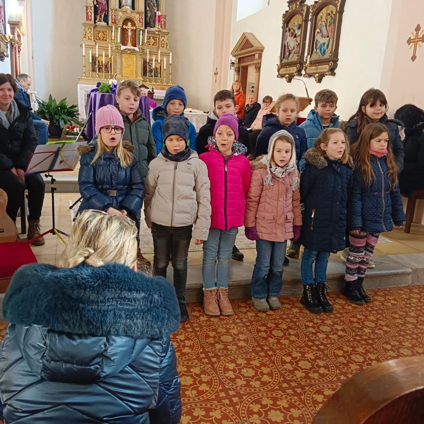 Eine Gruppe von Kindern in Winterjacken und Stiefeln singt in einer Kirche, mit einer Frau und einem Chor im Hintergrund.