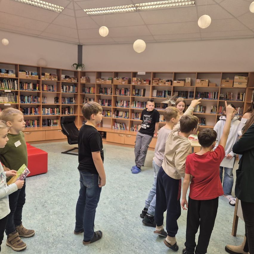 Eine Gruppe von Kindern steht in einer Bibliothek. Sie scheinen an einer Aktivität beteiligt zu sein. Hinter ihnen befinden sich Regale voller Bücher.
