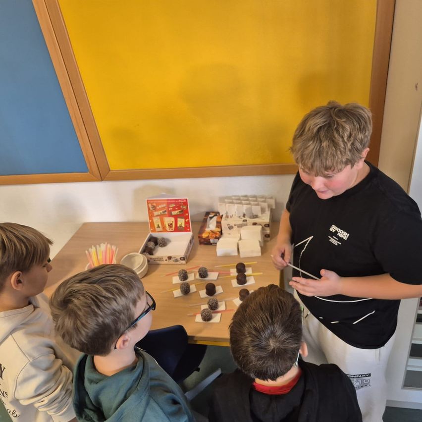 Ein Junge bereitet Schokolade auf einem Tisch zu, umgeben von anderen Jungen, die zusehen. Eine Schachtel Schokolade und eine gelbe Wand sind im Hintergrund.