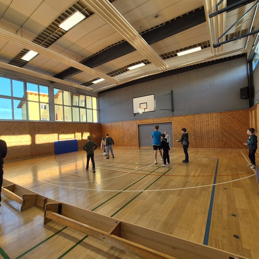 Eine Gruppe von Menschen spielt auf einem Hallenplatz. Sie stehen und unterhalten sich miteinander. Der Platz hat Holzböden und einen Basketballkorb.