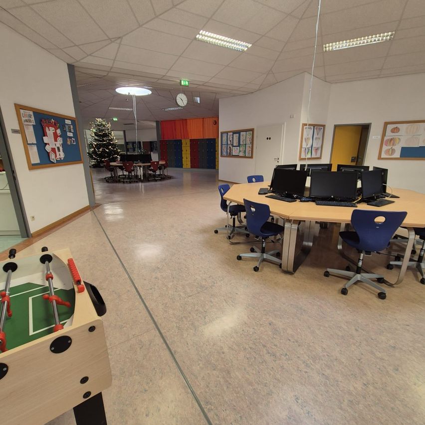 Ein leerer Klassenraum mit einem runden Tisch in der Mitte, umgeben von blauen Stühlen. Ein Tischfußball steht links. Ein Weihnachtsbaum ist im Hintergrund.