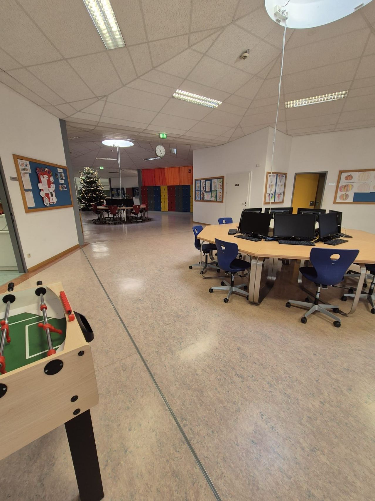 Ein leerer Klassenraum mit einem runden Tisch in der Mitte, umgeben von blauen Stühlen. Ein Tischfußball steht links. Ein Weihnachtsbaum ist im Hintergrund.