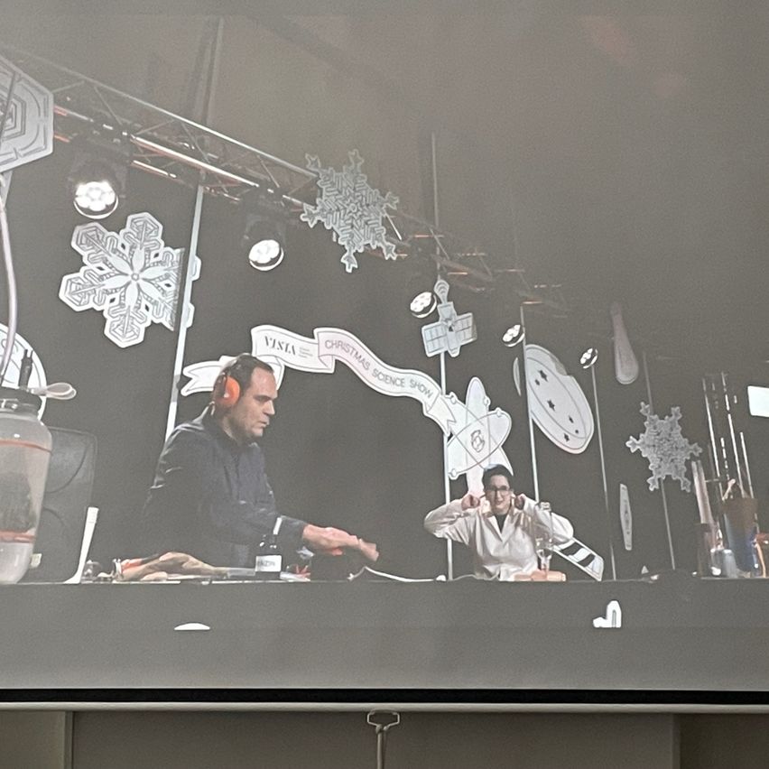 Zwei Personen sind auf der Bühne, eine trägt Kopfhörer, wahrscheinlich präsentieren sie eine Wissenschaftsshow. Der Hintergrund ist mit Schneeflocken und Weihnachtsdekorationen geschmückt.