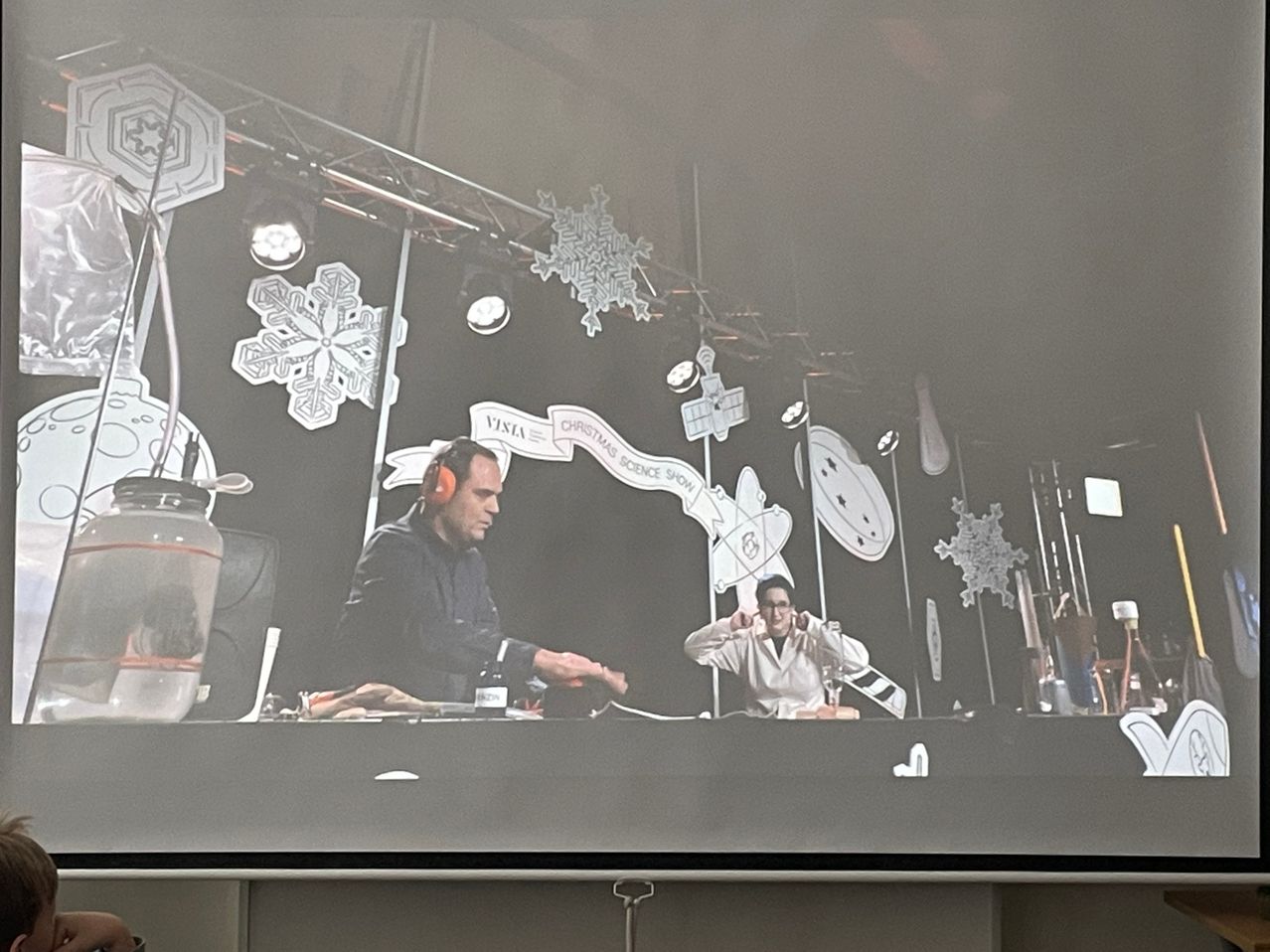 Zwei Personen sind auf der Bühne, eine trägt Kopfhörer, wahrscheinlich präsentieren sie eine Wissenschaftsshow. Der Hintergrund ist mit Schneeflocken und Weihnachtsdekorationen geschmückt.