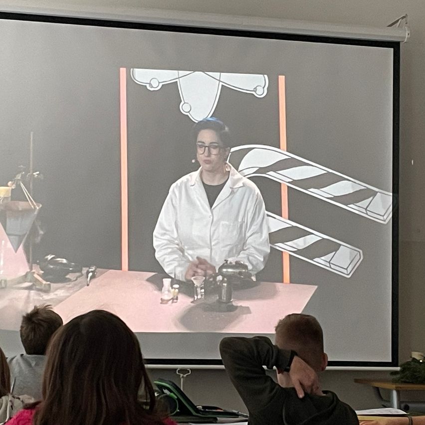 Eine Person in einem weißen Laborkittel und mit Brille wird auf einer Leinwand in einem Klassenzimmer projiziert. Mehrere Schüler schauen aufmerksam zu.