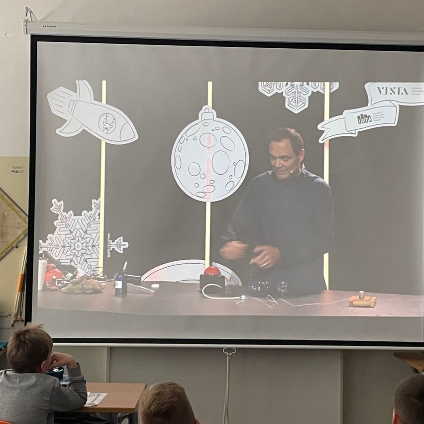Ein Mann präsentiert eine Dia-Show auf einer Projektionsleinwand mit Bildern von Rakete, Schneeflocke und Mond. Drei Kinder beobachten die Präsentation.