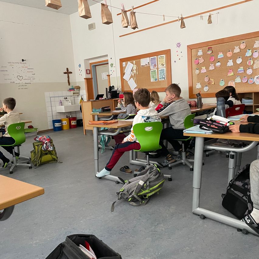 Ein Klassenzimmer mit Schülern, die an Tischen sitzen. Rucksäcke und Taschen liegen auf dem Boden. Pinnwände sind an den Wänden angebracht.