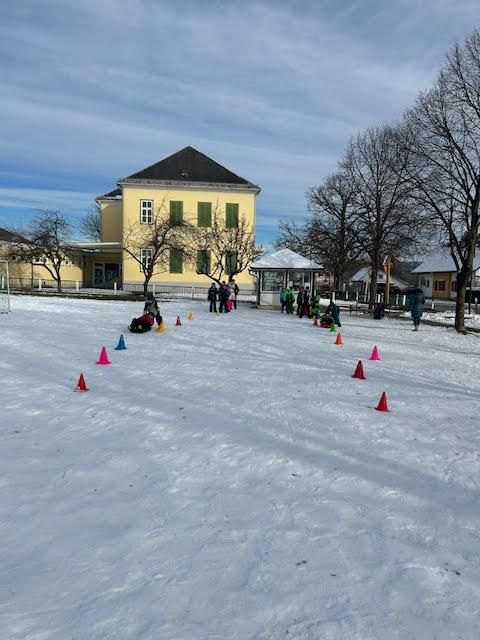 Eine Gruppe von Menschen spielt im Schnee mit einem gelben Gebäude und Bäumen im Hintergrund.