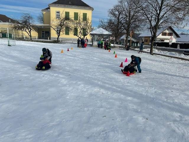 Kinder rodeln den verschneiten Hang hinunter, orangefarbene Kegel markieren den Weg, ein Gebäude im Hintergrund.