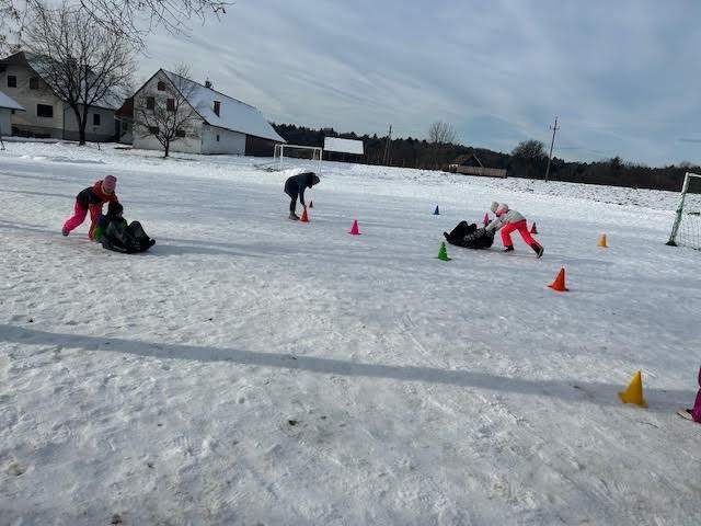 Kinder spielen auf einem verschneiten Feld mit Verkehrskegel. Ein Kind rutscht, während ein anderes hilft. Dahinter sind Häuser und ein Fußballtor zu sehen.