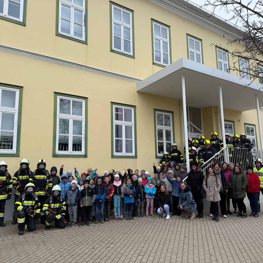 Eine Gruppe von Menschen, darunter Feuerwehrleute und Kinder, posiert für ein Foto vor einem gelben Gebäude.