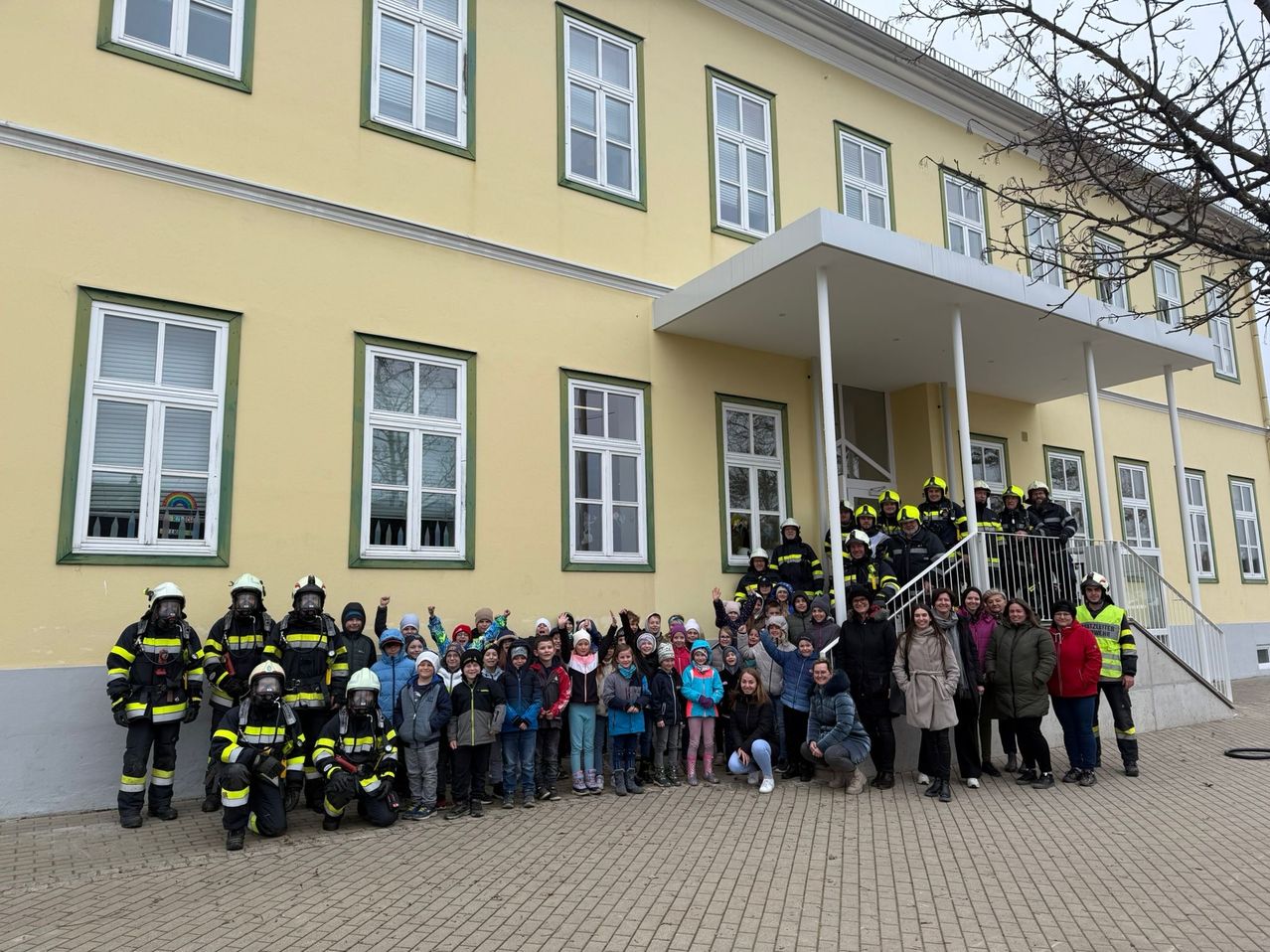 Eine Gruppe von Menschen, darunter Feuerwehrleute und Kinder, posiert für ein Foto vor einem gelben Gebäude.