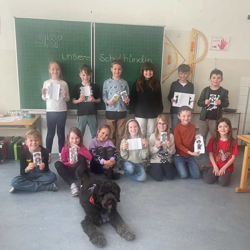 Eine Gruppe von Kindern und zwei Erwachsenen posiert für ein Foto in einem Klassenzimmer mit einem Hund. Sie halten alle Papiere in der Hand und lächeln. Dahinter steht eine Tafel mit den Worten 'Unsere Schulhunde'.
