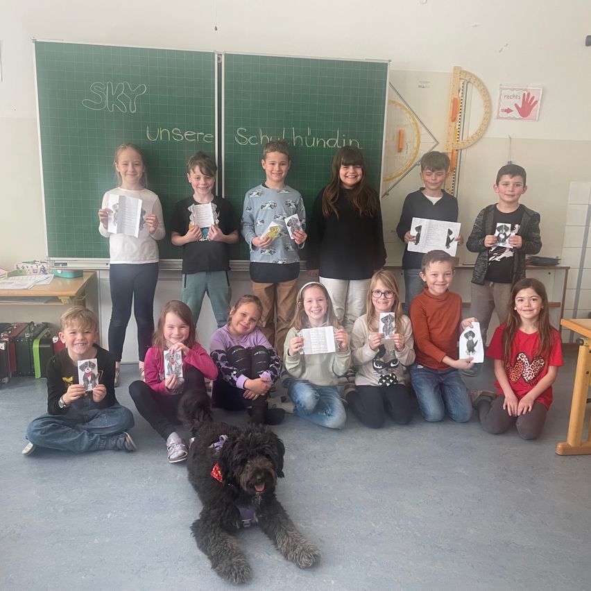 Eine Gruppe von Kindern und zwei Erwachsenen posiert für ein Foto in einem Klassenzimmer mit einem Hund. Sie halten alle Papiere in der Hand und lächeln. Dahinter steht eine Tafel mit den Worten 'Unsere Schulhunde'.