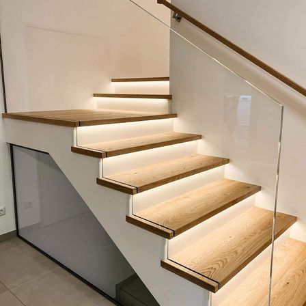 Moderne Treppe mit Holzstufen und LED-Beleuchtung. Klare Glasgeländer und ein hölzerne Handlauf. Weiße Wände und gefliester Boden.