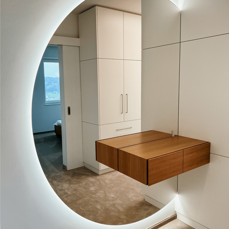 Ein minimalistischer Raum mit einer weißen Wand, einem Holzschrank und einem Spiegel. Der Boden ist mit Teppich ausgelegt und links befindet sich ein Fenster.