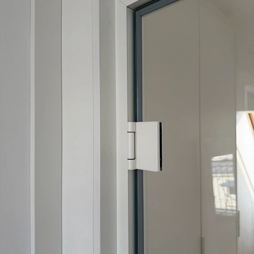 Nahaufnahme eines weißen Türscharniers, das an einer Glastür in einem Raum befestigt ist, mit einer Reflexion eines Balkons und eines Tisches.