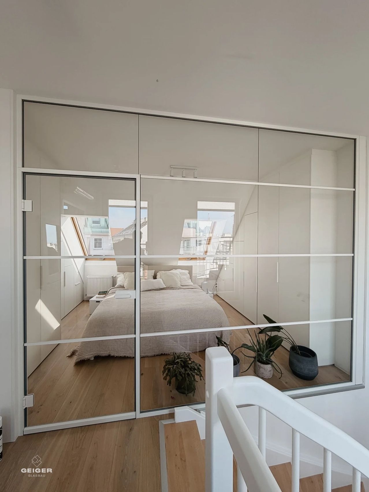 Ein Schlafzimmer mit Glaswänden und -türen, Holzboden, einem Bett mit Kissen, einem Nachttisch und Topfpflanzen.