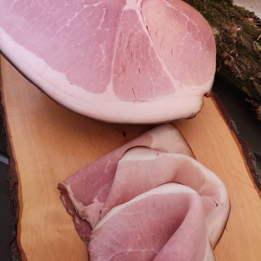 Ein ganzer Schinken und mehrere Schinkenscheiben auf einem Holzschneidbrett. Der Schinken hat eine glänzende rosa-rote Farbe und eine marmorierte Textur.