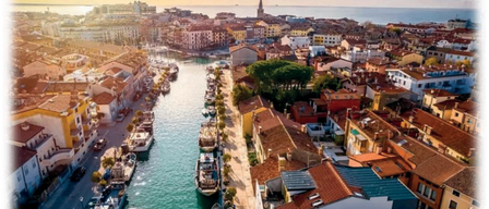 Luftaufnahme einer Küstenstadt mit an der Flussseite angedockten Booten, Gebäuden auf beiden Seiten und einem Turm in der Ferne.