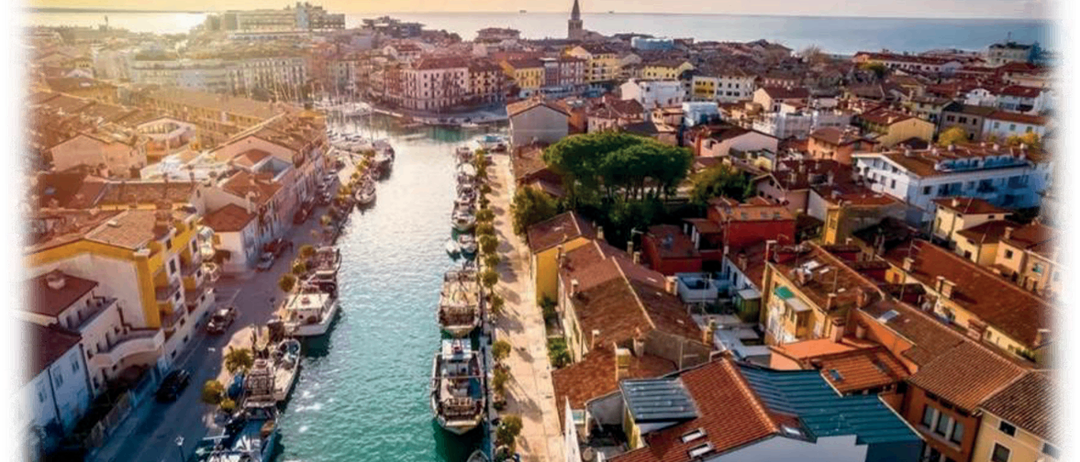 Luftaufnahme einer Küstenstadt mit an der Flussseite angedockten Booten, Gebäuden auf beiden Seiten und einem Turm in der Ferne.
