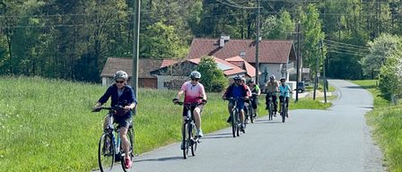Eine Gruppe von Radfahrern fährt eine Straße hinunter mit Häusern und Bäumen im Hintergrund. Die Radfahrer tragen Helme und fahren in einer Reihe.